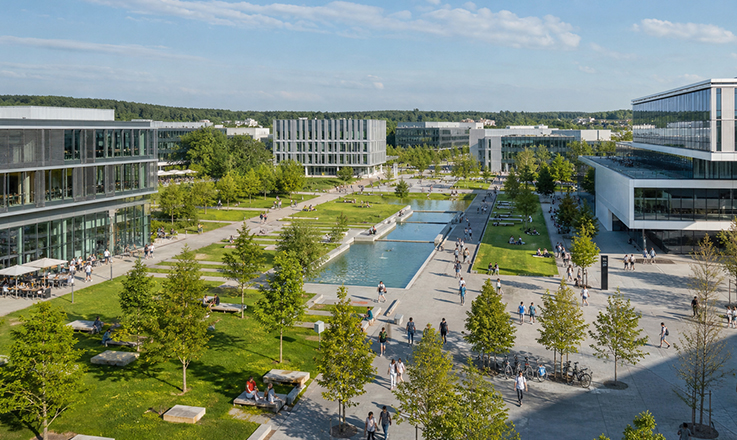 verweist auf: Chancen privater Hochschulen besser nutzen Moderne Campuslandschaft mit Wasserbecken, Grünanlagen, Fußwegen und Glasgebäuden; Menschen unterwegs an einem sonnigen Tag. (verweist auf: Chancen privater Hochschulen besser nutzen)